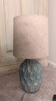 Full side view of the light green ceramic lamp with beige fabric shade on the carpeted floor near a wall base.