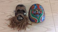 Photo of two masks facing forward showing front details: dark wooden mask with natural fiber beard and colorful Mexican style mask with bright patterns.