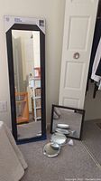 Photo shows one full-length black-framed mirror in packaging, one medium rectangular black-framed mirror leaning against a door, and three small round white-framed mirrors placed on the floor.