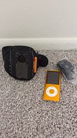 Orange Apple iPod Nano positioned upright with white click wheel visible, black adjustable armband beside it, earbuds in plastic wrapping placed nearby on carpet