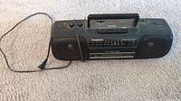 Photo of black Panasonic portable radio with AM/FM tuner, cassette player, buttons and knobs, carrying handle, and attached power cord, placed on carpet.