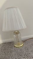 Small glass table lamp with decorative base and white pleated shade sitting on carpeted floor near wall.