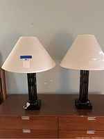 Two matching dark brownish-black lamps with off-white fabric shades placed side by side on a wooden dresser, showing overall condition.
