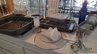 Display of various bakeware items including roasting pan, fish grilling baskets, cookie sheets and bread pans arranged on a counter.