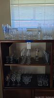 Full view of bar glass collection arranged on wooden shelves under window lighting. Includes varied styles of clear and patterned glasses.