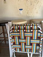 Two white wooden barstools and one black leather barstool with wood legs with padded round seats placed behind two colorful woven fabric folding chairs with white tubular metal frames.