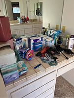 Bathroom counter showing various boxes of feminine hygiene products, tissues, personal care items, and electrical grooming tools including hair dryers and razors.