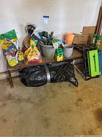 Wide shot showing the entire wooden garden bench with assorted gardening equipment including soil bags, pots, spreader, sprays, and gloves piled across it.