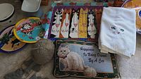 Photo showing assortment of cat-themed plates, cushion with embroidered cat, white towel with cat face embroidery, and a cat-shaped garden decoration.