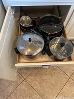 Drawer containing vintage aluminum Farberware cookware including pots, pans, and glass lids with black handles