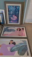 Three framed Hawaiian themed pictures leaning against wall, showing figures with tropical leaves