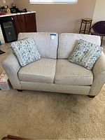 Front view of the tan fabric love seat with two patterned pillows on a carpeted floor near a tiled floor and kitchen area.