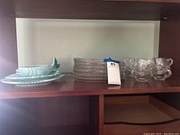 Shelf with seven clear glass luncheon plates, eight clear glass tea cups.