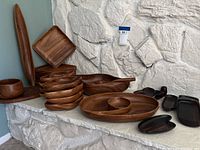 Photo showing multiple teak bowls, plates, and serving dishes arranged against a white stone backdrop. Includes elongated, round, and uniquely shaped pieces.