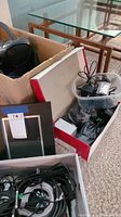 Box of assorted cords, power adapters, and electronics in open shoeboxes including a black device and cables
