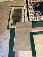 Overview of five rugs overlapping on tiled floor showing size, color, and pattern differences including a fringed floral green rug and solid green bordered rugs.