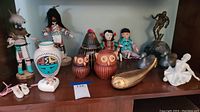Wide view of wooden kachina dolls, ceramic owl pottery, vase, metal fish sculpture, ceramic figurines, and other assorted decor placed on a wooden shelf.