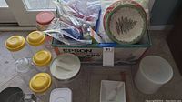 Various plastic jars with yellow lids, a white container, Christmas-themed plates, and other kitchen items in a box and on the floor