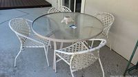 Round glass top patio table with four white woven chairs around it on concrete patio surface next to house wall.