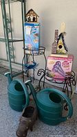 Photo of two green watering cans, two metal plant stands, and birdhouses placed on stands.