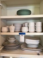 Photo showing white china dishes stacked on shelves, including plates, cups, bowls and casserole dishes.