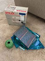 Singer Tiny Tailor sewing machine box, two folded fabric swatches, and green embroidery thread spool on carpeted floor.