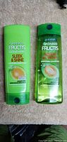 Photo of two green Garnier Fructis hair care products placed side by side on a beige carpet background. One is a squeeze tube conditioner, the other is a shampoo bottle.
