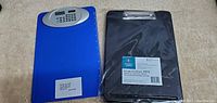 Two plastic clipboards, one blue with calculator and one black in packaging, on carpeted floor.