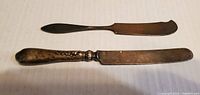 Two vintage silver butter knives displayed on a white surface. One has an ornate handle, the other a plain handle.