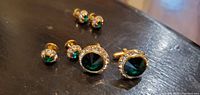 Photo showing two larger green jewel cufflinks with rhinestone borders and four smaller pairs with green jewels on a dark surface