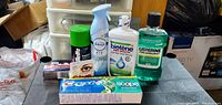 Photo shows seven hygiene and personal care items on a black surface including toothpaste, mouthwash, deodorant, air freshener, eye drops and lip care item.