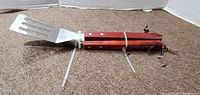 Photo of BBQ utensils showing spatula with wooden handle and slotted metal head, utensils tied together with zip tie.