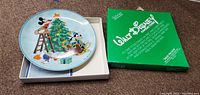 Photo of the collectible plate showing Mickey Mouse and Goofy decorating a Christmas tree with a green Disney-branded box beside it.