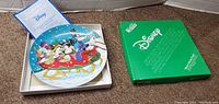Open box showcasing the collectible plate featuring Disney characters Mickey Mouse, Minnie Mouse, Donald Duck, and Goofy riding a sled in a snowy scene, along with the certificate of authenticity.