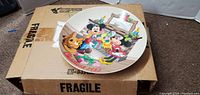 Top view of Disney collectible plate with Christmas 1986 design placed on a cardboard box labeled Fragile
