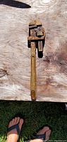 Photo of the full pipe wrench showing rusted metallic surface and wooden background, with part of a person's foot in a sandal visible at the bottom.