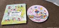 Photo of the collectible plate next to its colorful original box showing Disney characters on the box artwork.