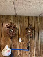 Photo showing two German Black Forest style cuckoo clocks hanging on wood panel wall, a larger one with stag head and smaller one with birds.