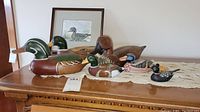 Photo showing a collection of decoy ducks on a lace table runner, including a large wooden mallard decoy, smaller painted ducks of varied species, and a framed duck print in the background.