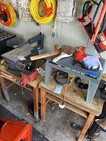 Wide view showing Sears Craftsman scroll saw on a wooden workbench, broom, sanding block, protective eyewear, power cords, and Hirsh Router & Sabre Saw Table with Black & Decker sabre saw on top.