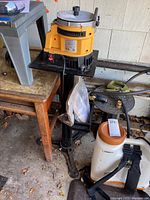 Photo showing the Work Sharp 2000 tool sharpener mounted on a black stand beside plastic and metal objects in a garage setting.