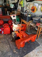 Side view of orange and white Simplicity gas snowblower near red fuel containers in workshop setting.
