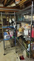 Photo showing two metal rolling wire storage racks with chrome finish, various household items on shelves, located in basement setting with concrete floor and exposed wooden ceiling.