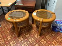 Pair of round wooden end tables with glass tops over woven plastic surfaces shown on patterned floor.