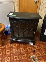 Front and slight angle view of the black electric fireplace stove, showing glass front with metal grid design and power cord.