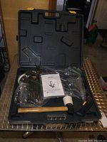 Photo of the carrying case opened showing the air powered nailer in plastic packaging, wooden mallet, and instruction manual sitting inside case compartments.