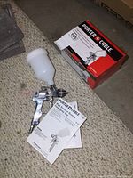 Two Porter Cable paint spray guns, one out of box and one near closed box with manual on carpeted floor.