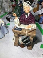 Front view of Royal Doulton Schoolmarm figurine seated at school desk with book, quill, and satchel visible.