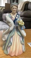 Full front view of the Royal Doulton porcelain figurine of Queen Elizabeth The Queen Mother dressed in a pastel gown with a blue sash and holding flowers and a handbag, showing detailed facial features and dress.