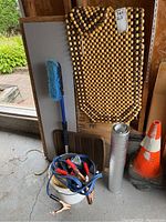 Photo showing jumper cables coiled next to black rubber car mats, a wooden bead seat mat hung on wall, a blue brush with handle, and foil-covered mat.